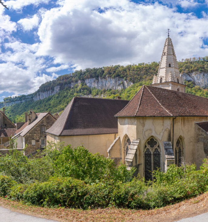 &Eacute;glise de village entour&eacute;e de verdure au pied d'une falaise sous un ciel nuageux.
