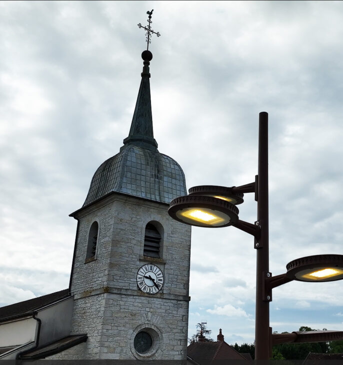 Clocher d'une &eacute;glise en pierre avec horloge et &eacute;clairage public sous un ciel nuageux.