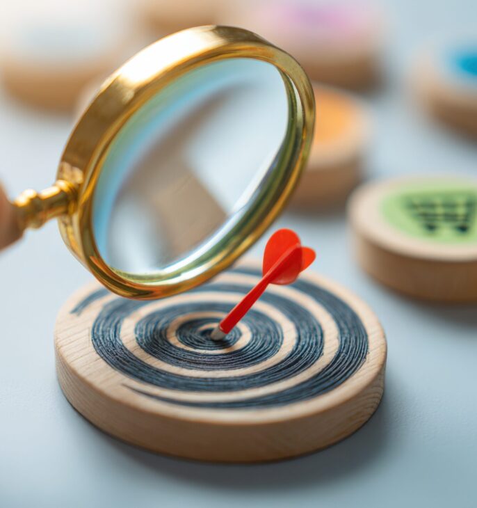main qui tiens une loupe sur une cible dans laquelle est plant&eacute; une fl&ecirc;chette le tout sur une pi&egrave;ce en bois et des caddie de diff&eacute;rents couleurs vives sont sur des pi&egrave;ces ronde en bois.